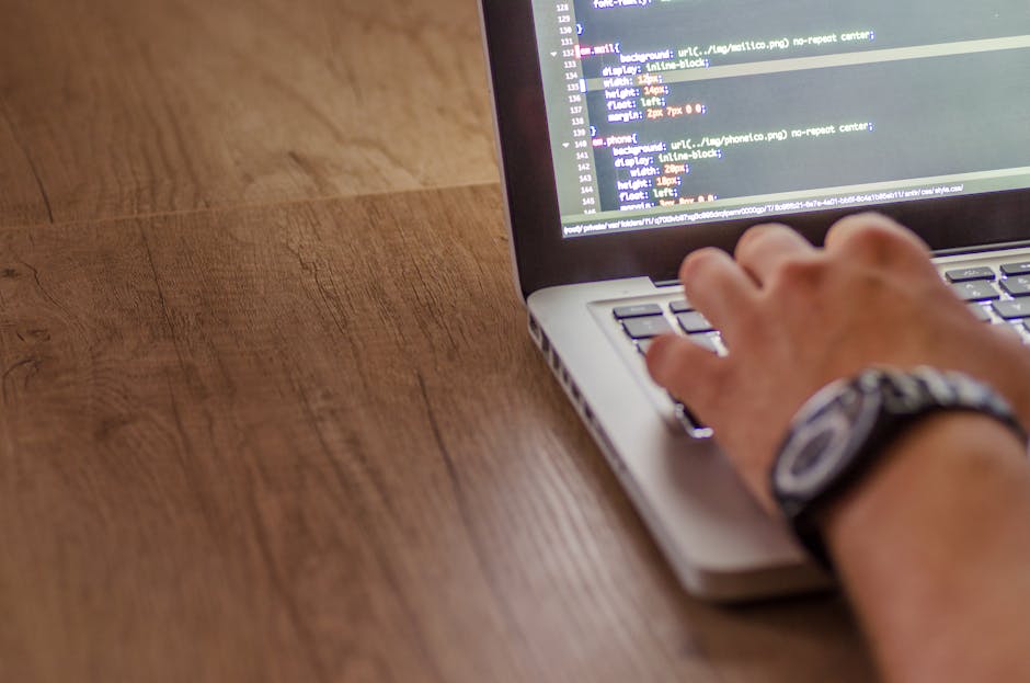 A person with a wristwatch is typing on a silver laptop placed on a wooden desk. The laptop screen displays a code editor with lines of programming code visible. The desk surface shows a smooth, polis