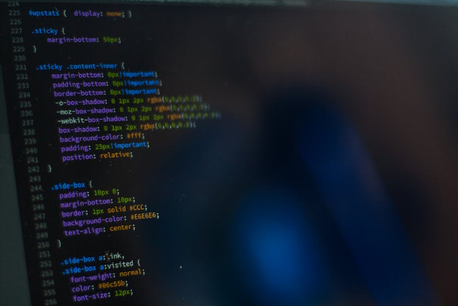 A person with a wristwatch is typing on a silver laptop placed on a wooden desk. The laptop screen displays a code editor with lines of programming code visible. The desk surface shows a smooth, polis