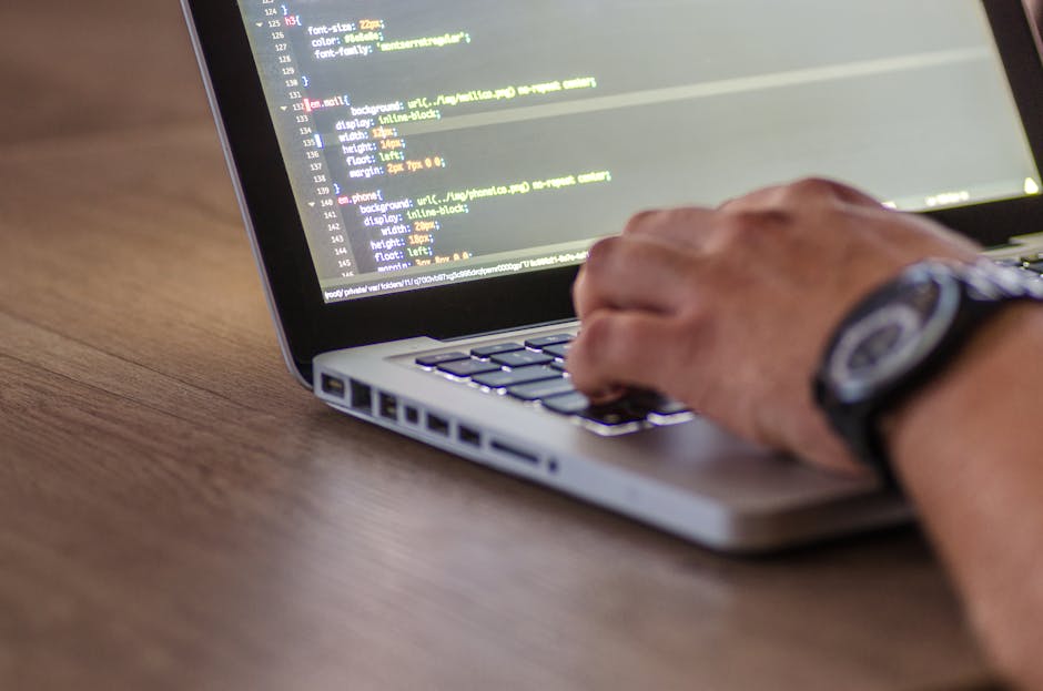 A close-up image of a person's hand typing on a silver laptop keyboard placed on a wooden desk. The laptop screen displays lines of HTML code with a dark background and multicolored text. The individu